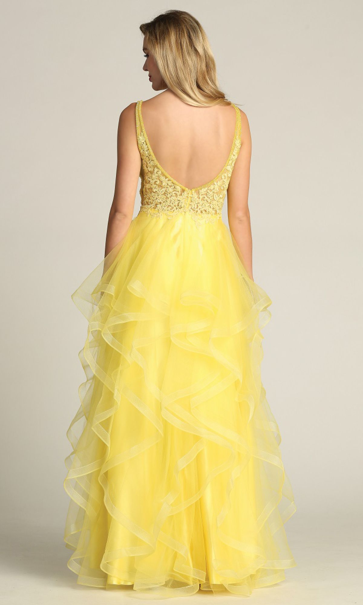 A woman stands facing away in the Sheer-Sides Long Tiered Prom Dress Chicas XR6006, featuring a sleeveless yellow beaded V-neck bodice, deep V-back, and layered tulle in an A-line silhouette.